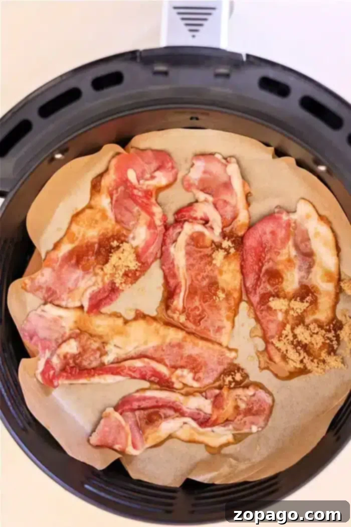 Air Fryer Bacon Candy 4 Slices of raw bacon, marbled with fat and kissed with maple syrup, are arranged on parchment paper inside an air fryer basket. The strips appear to be sprinkled with brown sugar, creating the perfect base for candied bacon.
