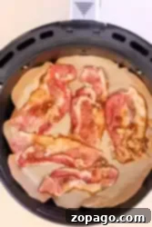 Slices of raw bacon, marbled with fat and kissed with maple syrup, are arranged on parchment paper inside an air fryer basket. The strips appear to be sprinkled with brown sugar, creating the perfect base for candied bacon.