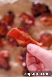 A hand holding a crispy piece of Air Fryer Maple Candied Bacon, with more maple-glazed bacon pieces blurred in the background.
