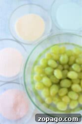 A bowl of green grapes, waiting to be transformed into candied delights with Jello, is surrounded by bowls of white, pink, peach, and cream-colored powders on a marble surface.