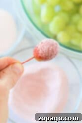 A hand holding a grape coated in pink sugary powder, showcasing how to make candied grapes with Jello; green grapes are visible in a bowl in the background.