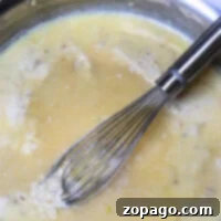 A whisk in a pan gently stirs the creamy mixture with bits of food, showing the magic of how to make gravy from chicken drippings, all on a pristine white surface.