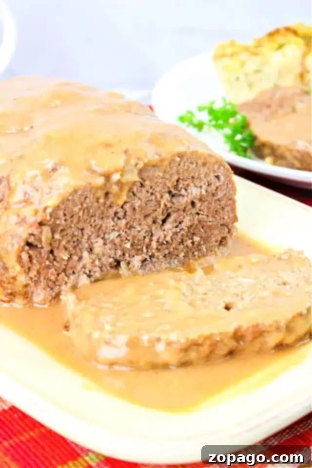 Ultimate Meatloaf Gravy 2 A perfectly sliced loaf of meatloaf, generously topped with glistening brown gravy, rests on a pristine white plate. The rich, dark gravy cascades over the edges, promising a burst of savory flavor. In the soft-focus background, side dishes like vibrant green peas complete this enticing meal, emphasizing the perfect pairing of our homemade brown gravy with a classic meatloaf recipe.