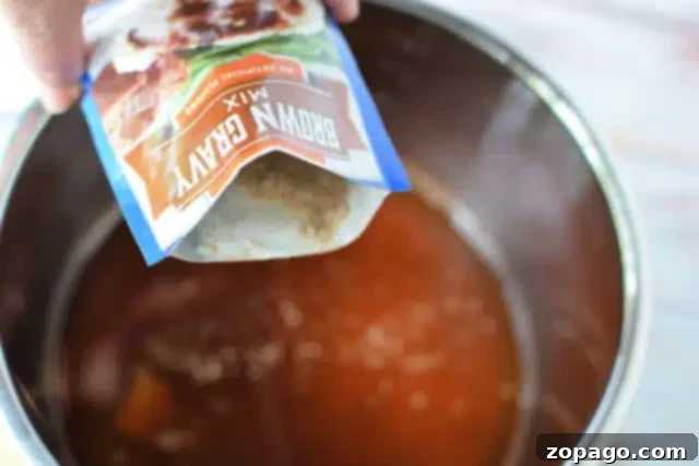 Ultimate Meatloaf Gravy 3 A hand is pouring a packet of brown gravy for meatloaf recipe mix into a metal pot filled with liquid. The contents are partially visible as they blend into the pot.