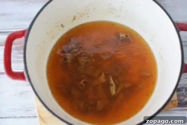 Ultimate Meatloaf Gravy 4 A red-handled pot containing simmering broth reminiscent of a brown gravy for meatloaf recipe with visible meat bones rests on a wooden surface. The rich, brown broth is slightly foamy at the edges.