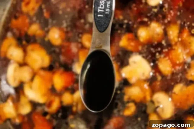 Ultimate Meatloaf Gravy 6 Close-up of a tablespoon labeled with 1 Tbsp, 15 mL, hovering above a pot of chickpeas, tomatoes, and spices. The spoon holds brown gravy, likely a rich seasoning for a meatloaf recipe.