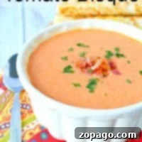 A bowl of creamy tomato bisque, crafted from an easy tomato bisque recipe, is garnished with herbs and sits beside a spoon on a colorful cloth.