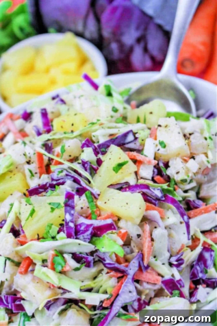 Creamy Hawaiian Pineapple Slaw 2 A vibrant bowl of Hawaiian Pineapple Coleslaw featuring purple cabbage, carrots, pineapple chunks, and fresh herbs, with a spoon on the side.