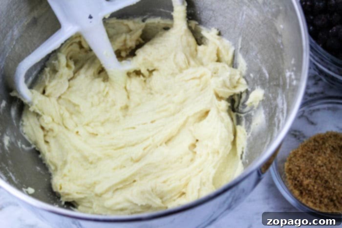 Muffin batter is being mixed in a metal bowl with a paddle attachment, surrounded by bowls of brown sugar and chocolate chips. The scene evokes the cozy ambiance of baking jumbo blueberry muffins.