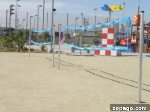 Multiple beach volleyball courts on a sandy ground with blue nets are located near an outdoor recreational area at Perris Water Park, featuring colorful structures like a red and white checkered tower and a blue airplane. Floodlights and poles stand visible in the background.