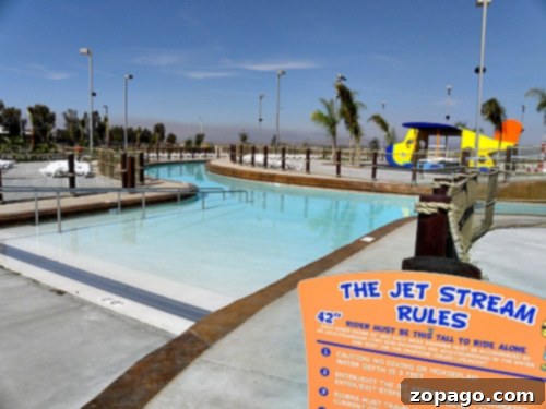 A water park with a winding lazy river and a colorful play structure in the background. The foreground features a sign labeled "The Jet Stream Rules" with text about height and safety requirements, showcasing the fun and safety standards at Perris Water Park.