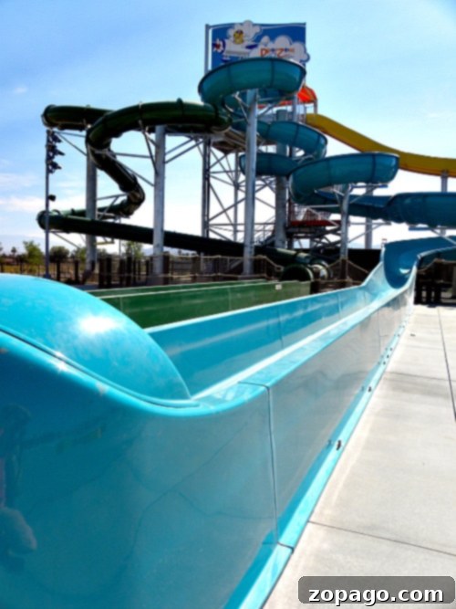 A large water slide structure at Perris Water Park features multiple spiraling tubes of green and yellow. The blue slide leads to a splashdown area on a concrete surface. A sign with a cartoon figure is visible at the top of the structure. The sky is clear and sunny.