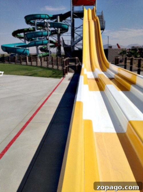 A view of Perris Water Park's slide area features multiple yellow and white slides side by side. In the background, a spiraling green and blue water slide structure is visible. The area around the slides is paved, and there is a fence separating the slides from the grass.
