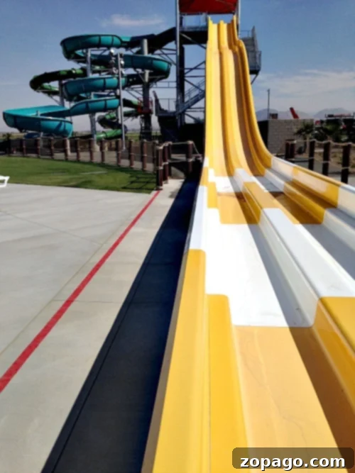 DropZone Water Park Perris What You Need To Know 6 A view of Perris Water Park's slide area features multiple yellow and white slides side by side. In the background, a spiraling green and blue water slide structure is visible. The area around the slides is paved, and there is a fence separating the slides from the grass.