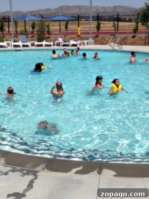 A group of people, including children wearing yellow floatation devices, swim and play in the pool at Perris Water Park. Lounge chairs are aligned along the poolside with a few people sitting and relaxing. A fenced area and a mountainous landscape are visible in the background.
