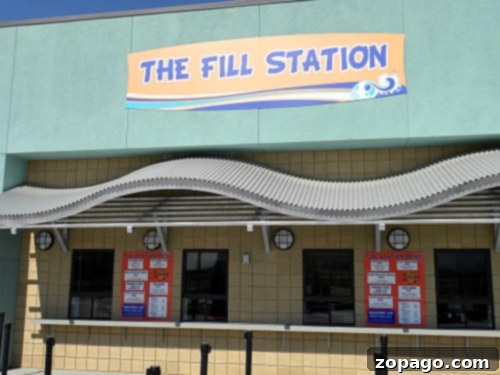 The image shows an establishment called "The Fill Station" with a sign above and a wave-like awning, reminiscent of Perris Water Park. Below are three service windows with red menus posted on the wall. The building has a light green and yellow exterior.