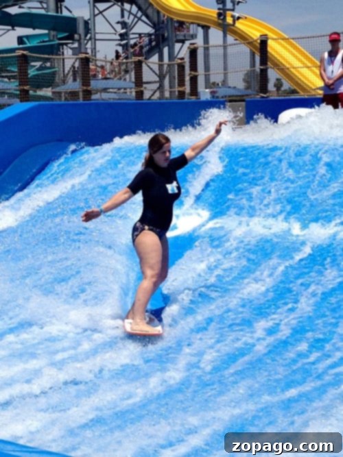 A person in a black wetsuit rides a wave on a bodyboard at Perris Water Park, maintaining balance with arms outstretched. A yellow water slide is visible in the background, along with several other people looking on.