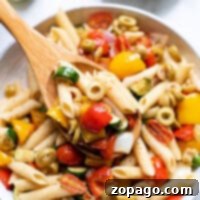 A bowl of roasted vegetable pasta salad with penne, tomatoes, olives, zucchini, and bell peppers, served with a wooden spoon.