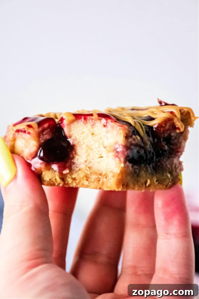 Close-up of a hand holding a delicious peanut butter and jelly cheesecake bar with a bite taken out, showing the layers.