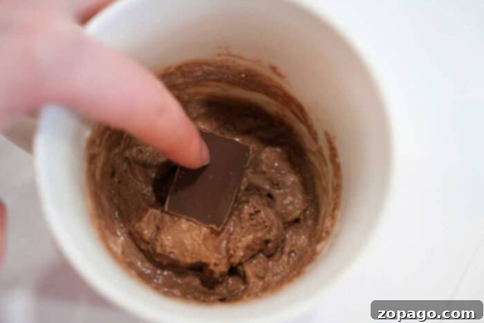 Mug Lava Cake Magic 4 Pressing a chocolate square into chocolate batter inside a white mug, preparing a quick and delicious Lava Cake.