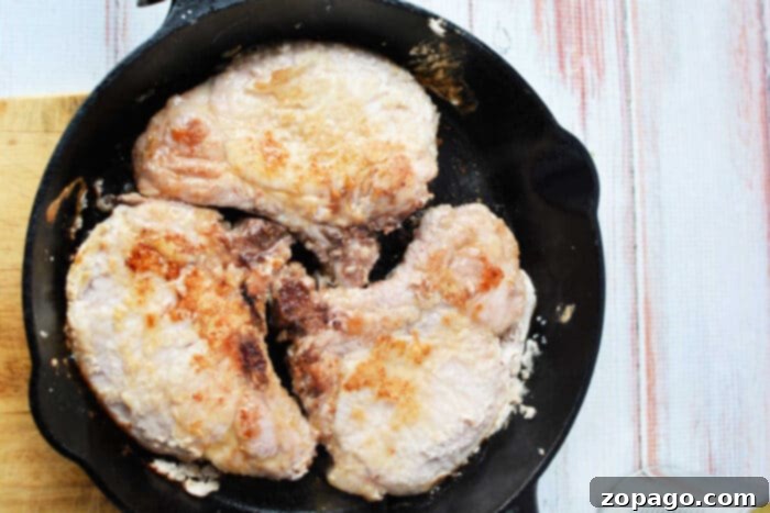 Four pork chops cooking in a black cast iron skillet on a light wooden surface, perfect for a savory pork and pineapple meal.