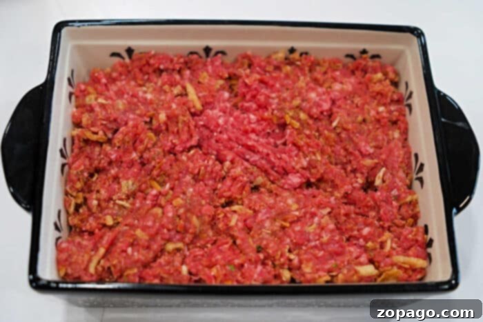 Smoky BBQ Meatloaf Perfection 5 Raw ground beef mixed with onions is spread evenly in a rectangular baking dish, perfect for a classic meatloaf recipe and ready for cooking.