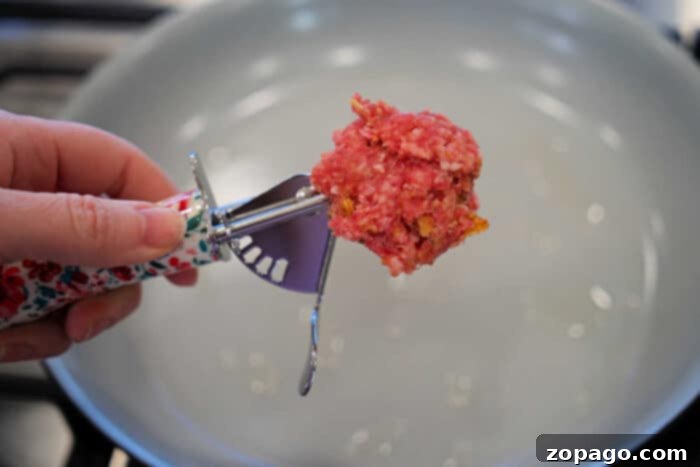Cheesy Baked Spaghetti Meatball Bake 4 Hand holding a scoop of raw ground meat over an empty frying pan on a stove, perfect for preparing baked spaghetti with meatballs.
