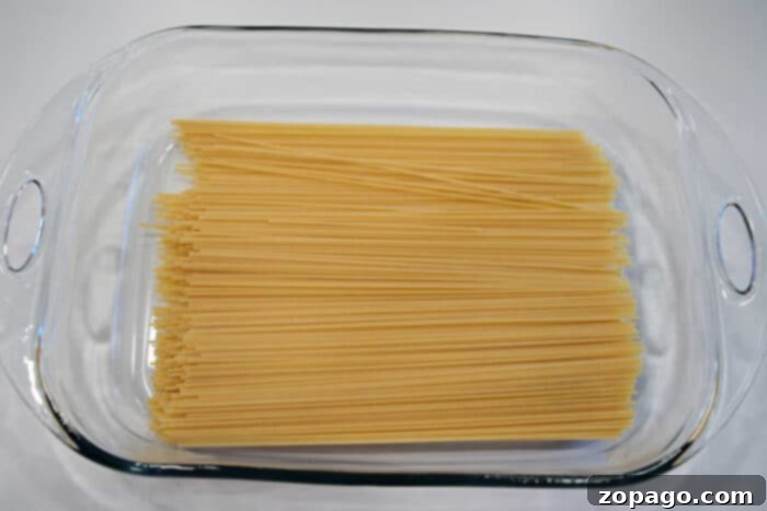 Cheesy Baked Spaghetti Meatball Bake 5 Uncooked spaghetti noodles arranged in a clear glass baking dish on a white surface, ready to be transformed into delicious baked spaghetti with meatballs.