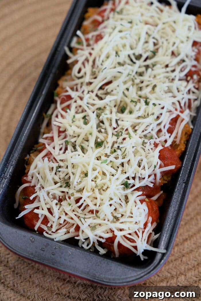 Chicken Parmesan Meatloaf Recipe That Melts in Your Mouth 4 A loaf pan filled with Chicken Parmesan Meatloaf, topped with shredded cheese and herbs, ready to be baked.
