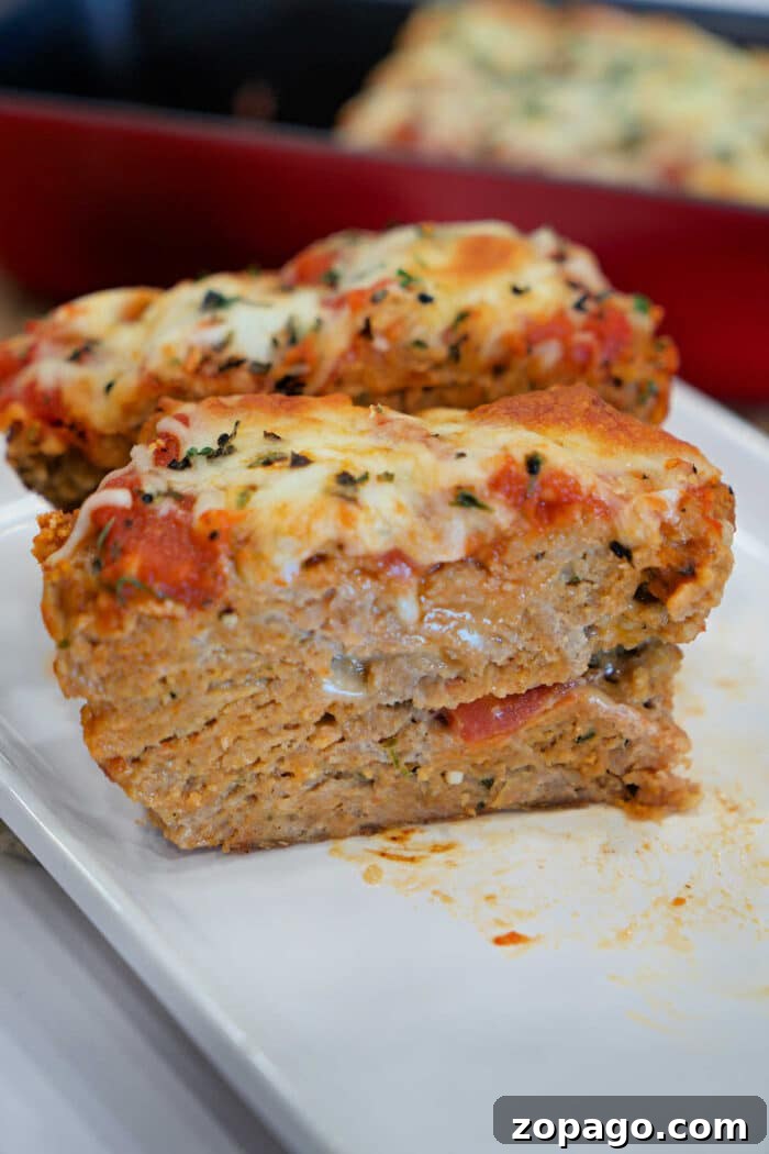 Chicken Parmesan Meatloaf Recipe That Melts in Your Mouth 5 Two slices of cheesy, baked Chicken Parmesan Meatloaf topped with tomato sauce and herbs sit on a white plate.