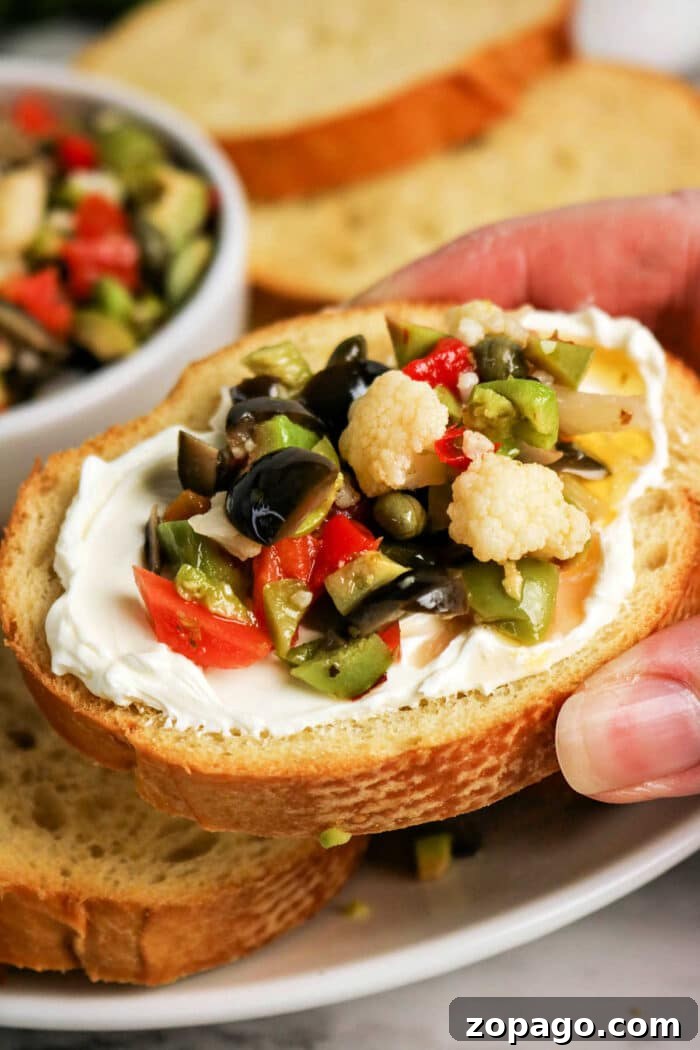 Authentic Muffuletta Olive Salad Recipe for Sandwichs & Platters 2 A hand holds a slice of bread topped with cream cheese and a vibrant Olive Salad, featuring chopped olives, cauliflower, and peppers—a perfect bite inspired by the classic Muffuletta recipe.
