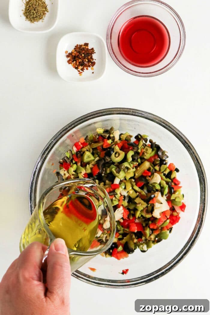 Authentic Muffuletta Olive Salad Recipe for Sandwichs & Platters 4 A hand pours olive oil into a bowl of chopped vegetables for a classic Olive Salad, with spices and red vinegar nearby—perfect for an authentic Muffuletta recipe.