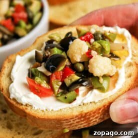 Authentic Muffuletta Olive Salad Recipe for Sandwichs & Platters 6 A hand holds a slice of bread topped with cream cheese and a vibrant Olive Salad, featuring chopped olives, cauliflower, and peppers—a perfect bite inspired by the classic Muffuletta recipe.