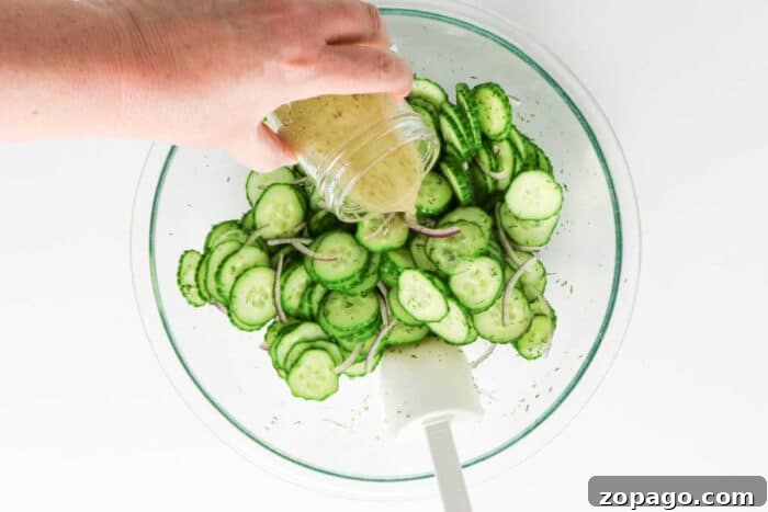 Cucumber and Dill Salad with Creamy Yogurt Dressing 5 A hand pours dressing over sliced cucumber and onion in a glass bowl with a white spatula, creating a fresh salad perfect for adding dill.