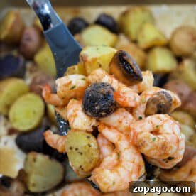 A spoon holds shrimp and potatoes with mushrooms above a tray filled with roasted shrimp and potatoes, plus savory mushrooms.