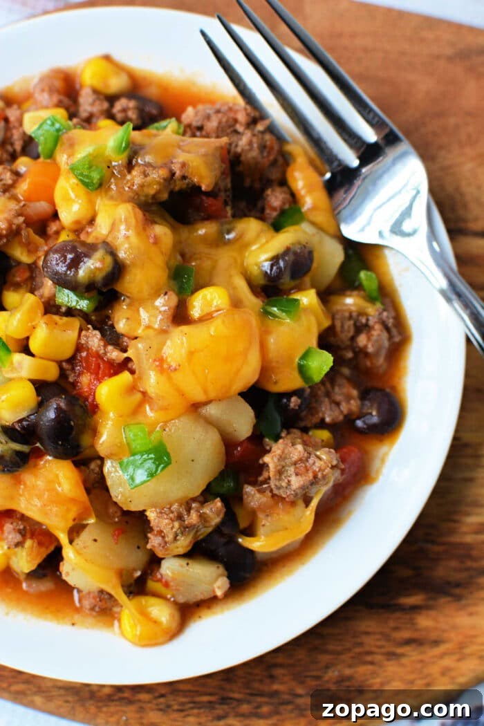Slow Cooker Cowboy Casserole 5 A plate of cheesy Cowboy Casserole with beef, black beans, corn, and potatoes sits next to a fork—perfect for fans of hearty Crockpot recipes.