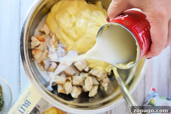 Cheesy Ritz Chicken Casserole with Crunchy Cracker Topping 4 A hand pours canned milk into a bowl with diced chicken and creamy yellow sauce, preparing a delicious mixture for Ritz Chicken Casserole.