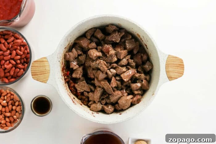 Slow-Simmered Chuck Roast Chili Recipe 5 Overhead view of cubed beef in a white pot, perfect for making Chuck Roast Chili, surrounded by beans, sauce, and seasonings on a white surface.