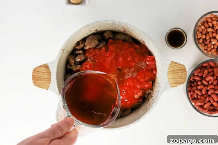 Slow-Simmered Chuck Roast Chili Recipe 6 A hand pours broth into a pot with beef and tomatoes, surrounded by beans and sauce on a white table, creating the perfect base for a hearty beef chili recipe.